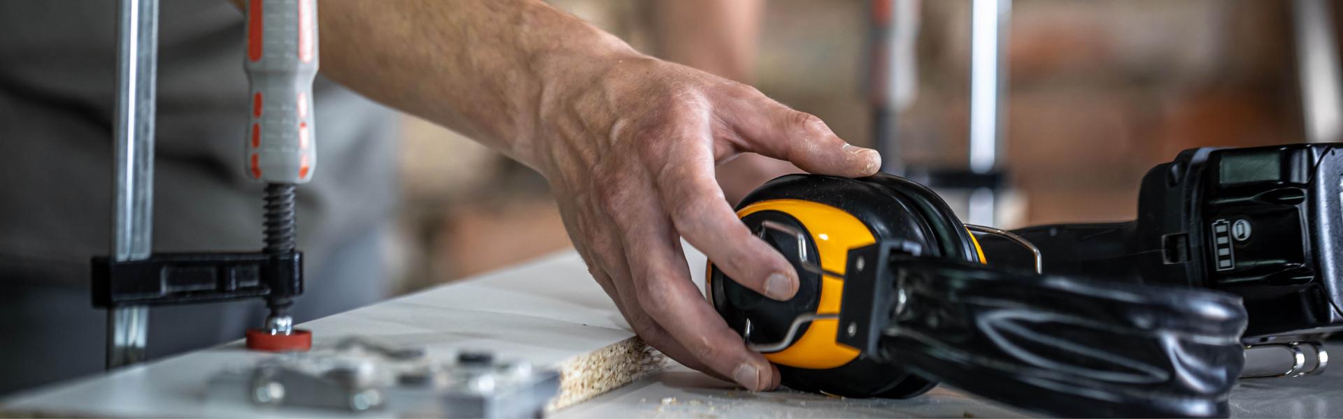 Worker's hand picking up full coverage earphones from a workbench with tools.